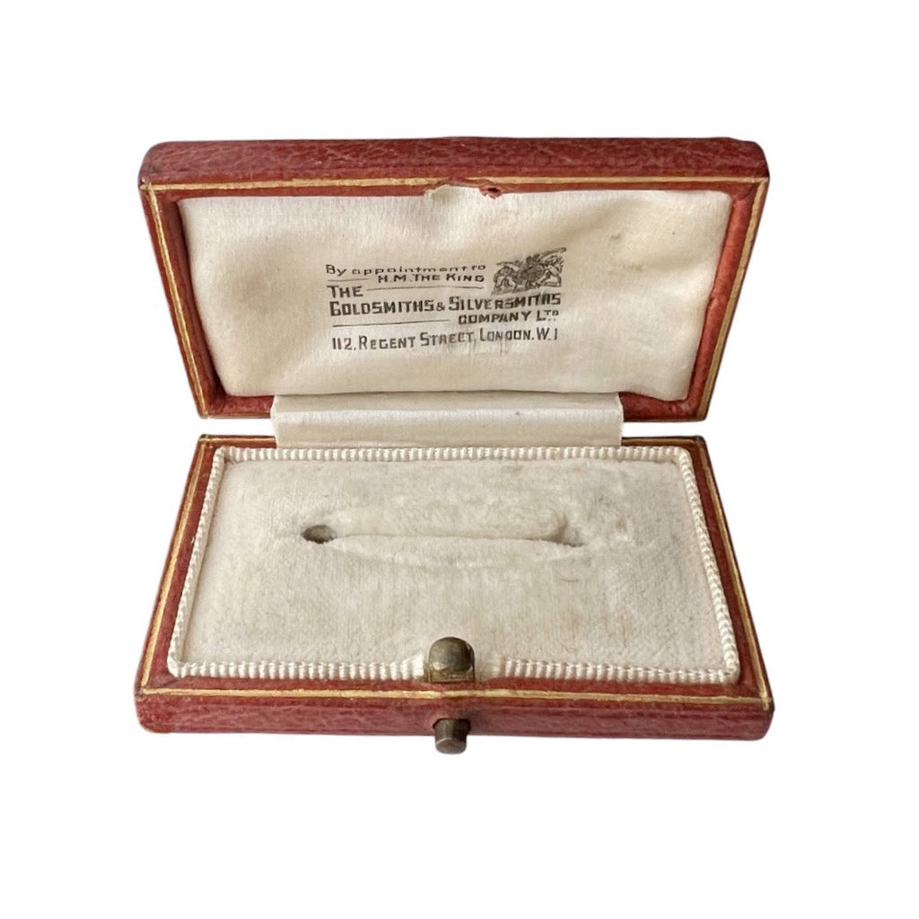 An open antique brooch box with a reddish-brown wood exterior and cream-colored velvet interior, displaying a small silver-colored object inside. The inside lid features printed text reading 'The Goldsmiths & Silvermith Co Ltd'.