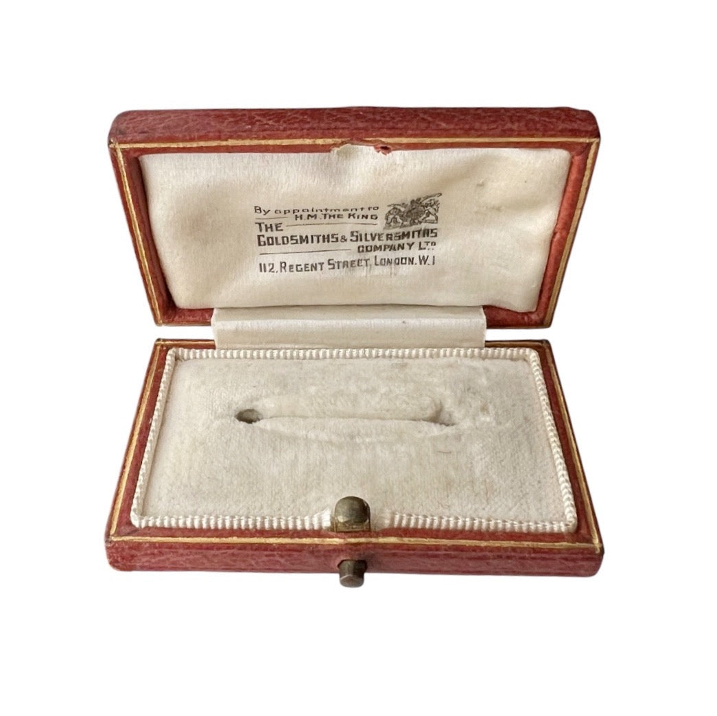 An open antique brooch box with a reddish-brown wood exterior and cream-colored velvet interior, displaying a small silver-colored object inside. The inside lid features printed text reading 'The Goldsmiths & Silvermith Co Ltd'.