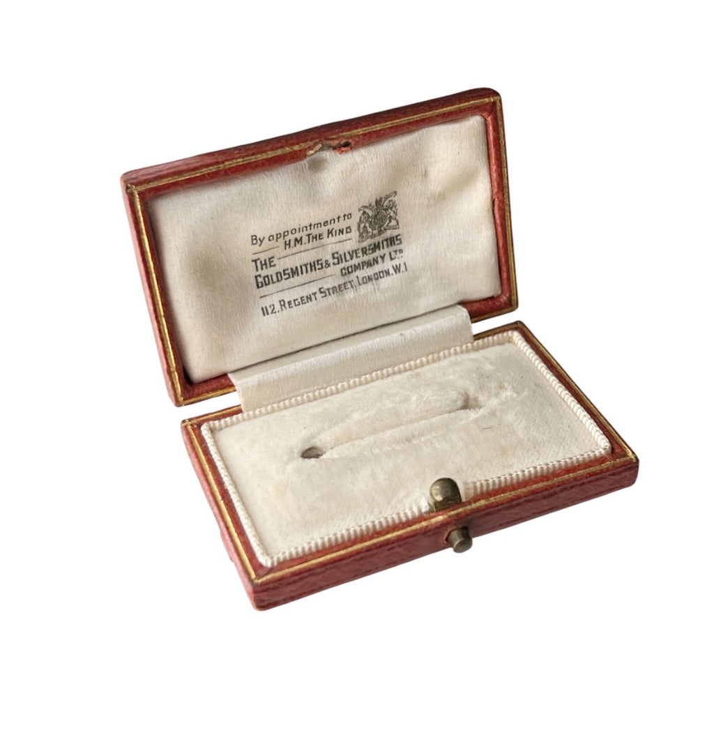 An open antique leather brooch box with cream velvet lining, displaying a silver pin and an interior label reading 'The Goldsmiths' Company.'.